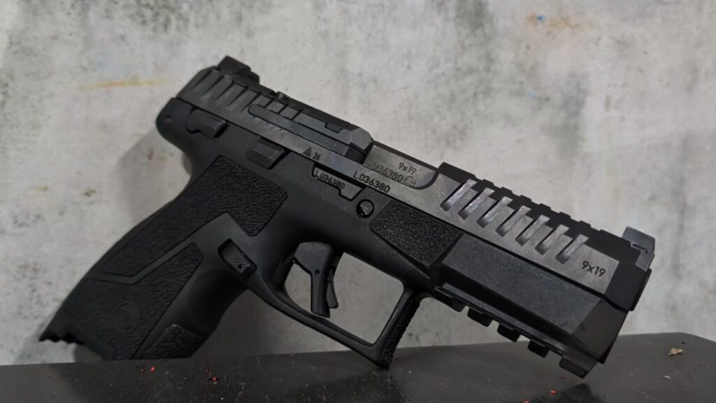 A close-up low-angle shot of a black CZ Duty 9mm pistol with standard iron sights resting on a dark surface against a mottled gray background.