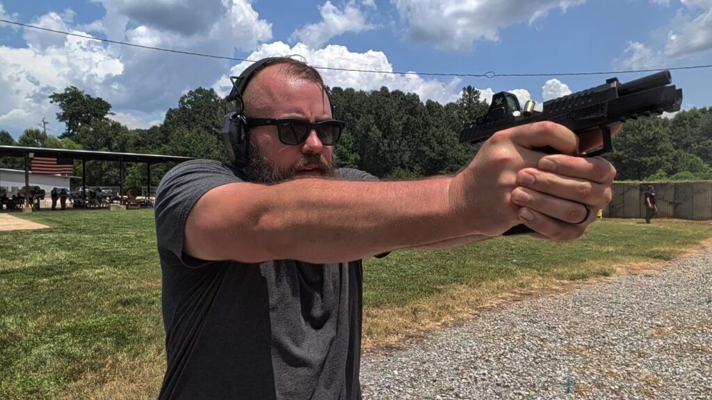 Bearded shooter in sunglasses and ear protection firing a CZ DUTY with a red dot, slide locked back, at an outdoor range