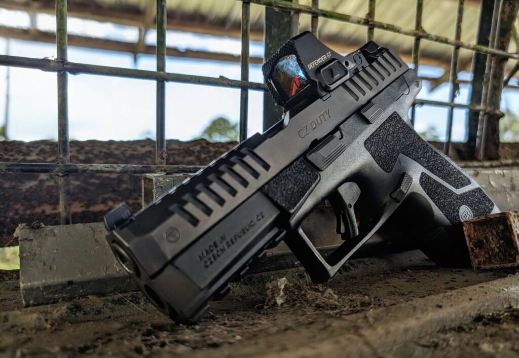 A low-angle view of a black CZ Duty pistol with a red dot sight resting on a rustic ledge in front of a wire fence