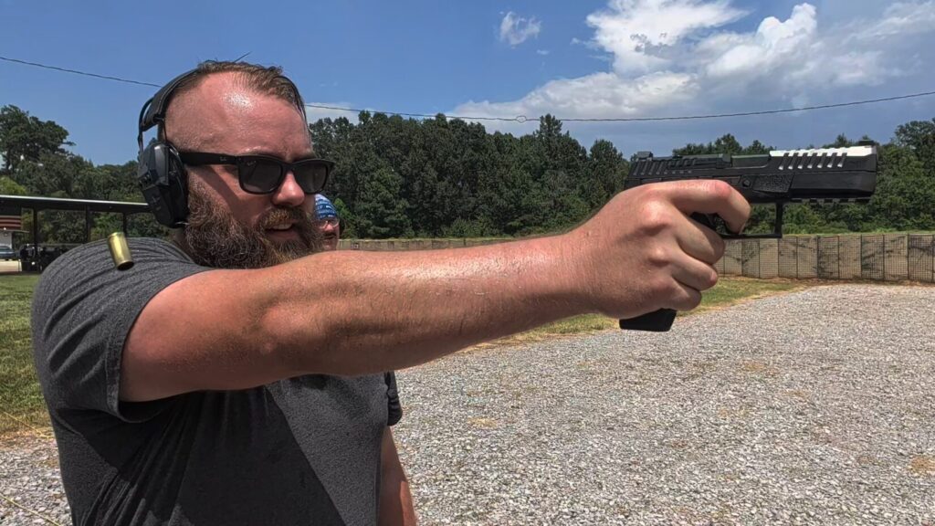 Travis Pike firing a CZ Duty pistol at an outdoor range, with a spent brass casing caught mid-air as it ejects from the firearm.